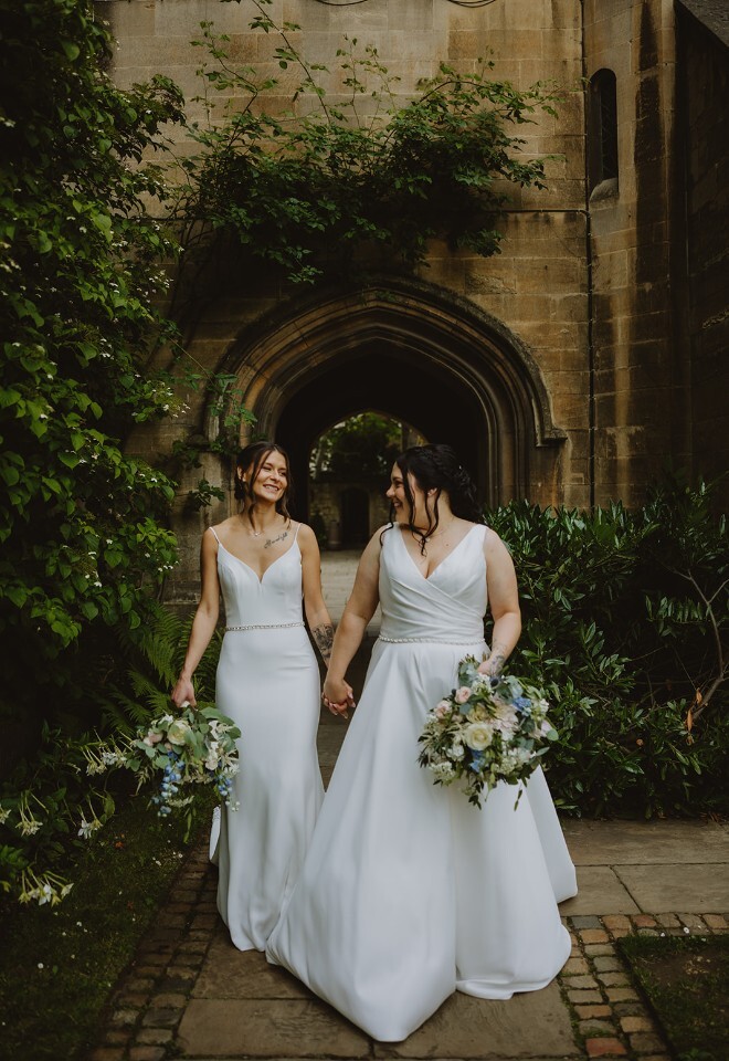 Brides holding hands and smiling as they walk through a historic Oxford cloister garden, photographed by V & H Photography.