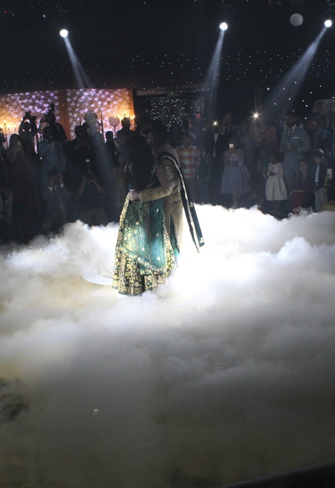 A couple in traditional South Asian attire embraces during a dance on a fog-covered floor, illuminated by bright spotlights while guests watch from a dimly lit venue.