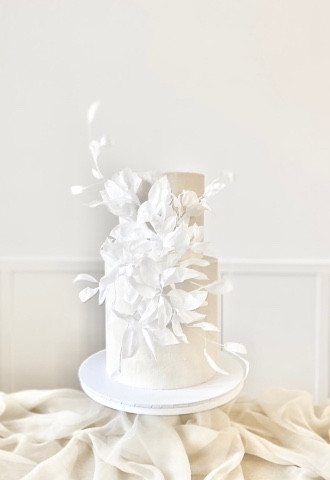 White two tier ethereal rice paper white wedding cake on a white cake stand above a bed of cream chiffon drape with a neutral background