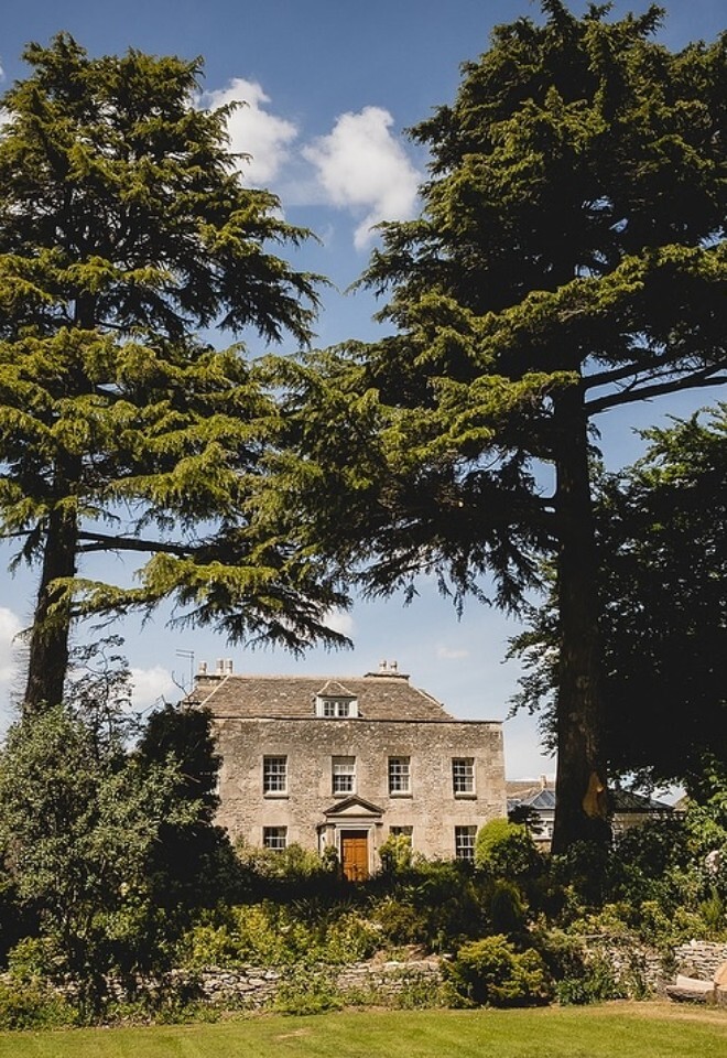 Historic stone manor house at Great Tythe Barn, framed by tall cedar trees and lush gardens under a bright blue sky, with sunlight illuminating the façade and surrounding greenery.