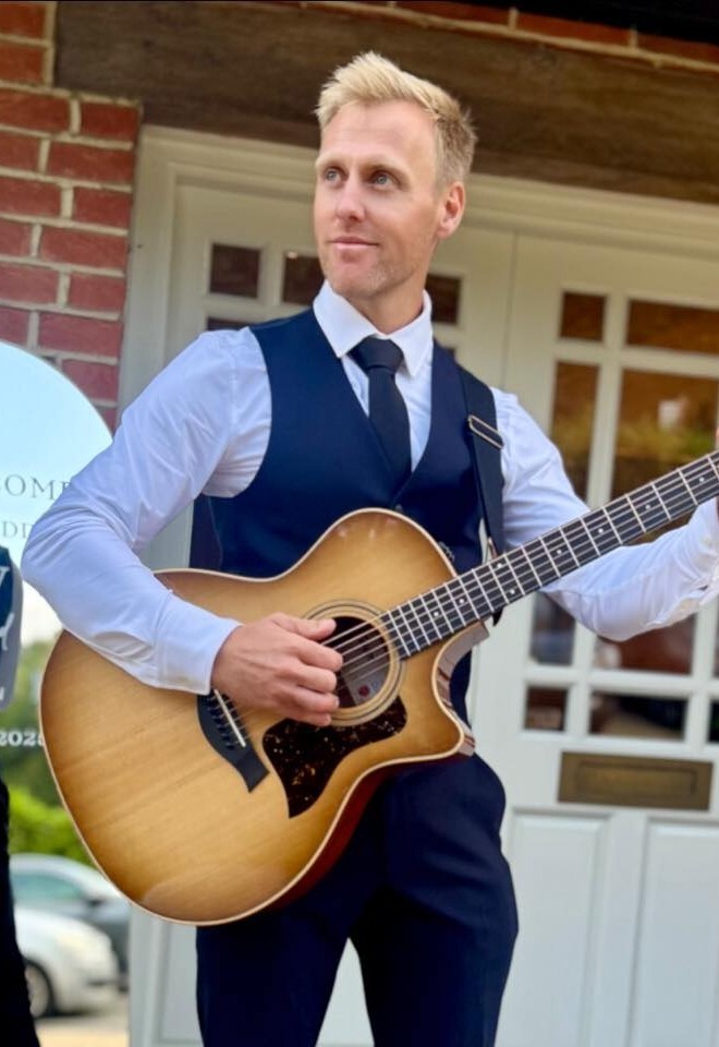 Matt Rayner, an Essex-based roaming wedding musician, plays acoustic guitar outside a wedding venue, dressed in a smart waistcoat and tie.