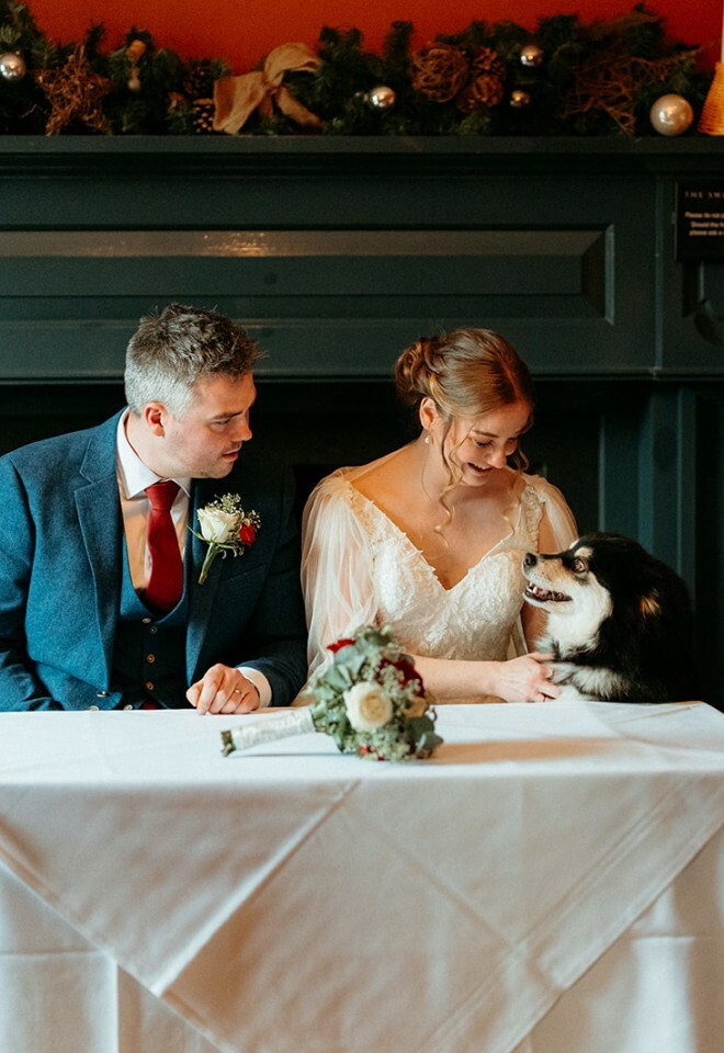 Bride and groom sit at registrar table looking down at black and white fluffy dog
