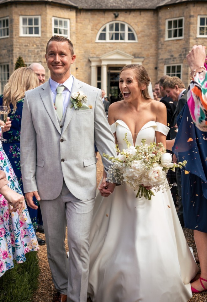 Bride and groom walk hand in hand outside Washingborough Hall Hotel, smiling as guests in colourful outfits throw confetti and celebrate their wedding day.