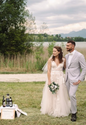 The officiant is standing in a meadow with the bride and groom, in between them is a table with two beers for a blending ceremony