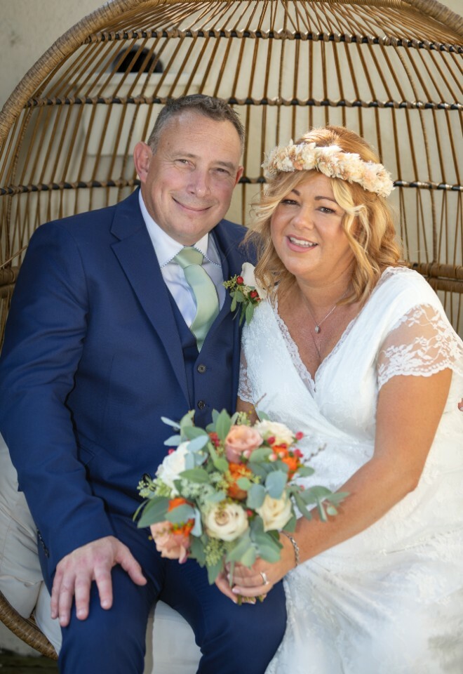 A bride and groom sit together in a wicker egg chair, with the bride holding a peach and cream floral bouquet.