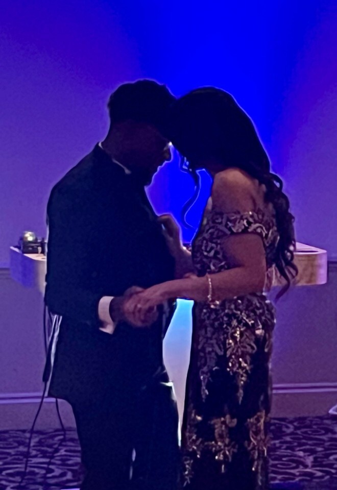 A couple leaning close together during their first dance at an Afro-Indian wedding reception, framed by vertical LED light bars under deep blue stage lighting.