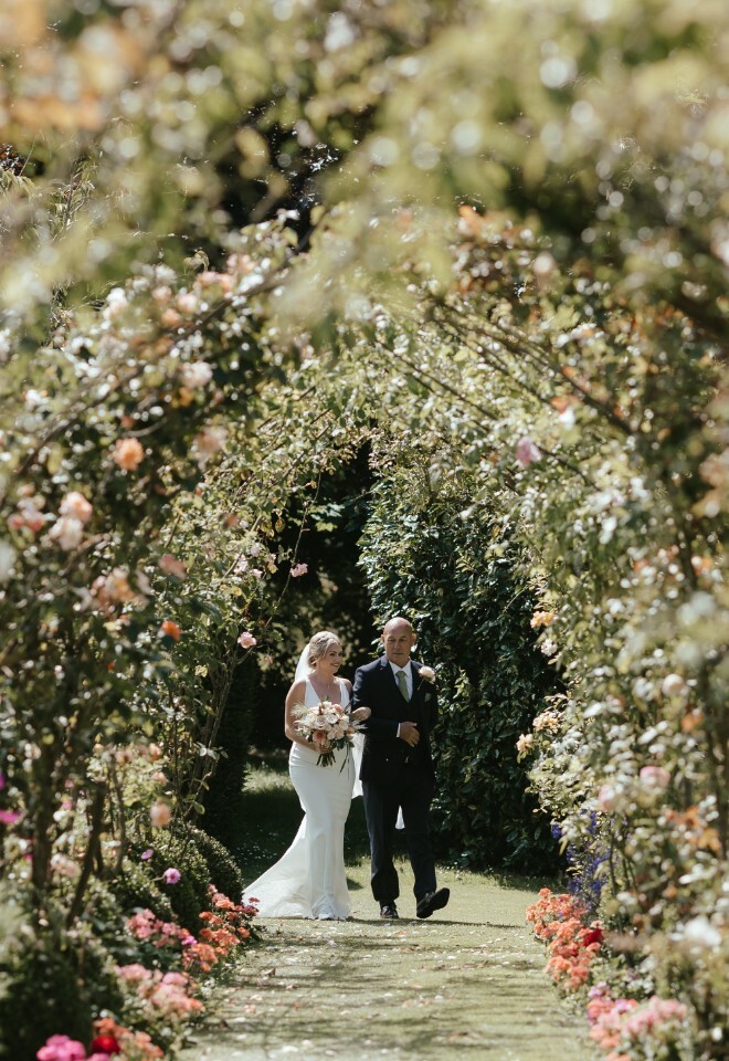 July Outdoor Wedding Ceremony, Bride Walking Down the Aisle under the Arch of Roses with Father of the Bride