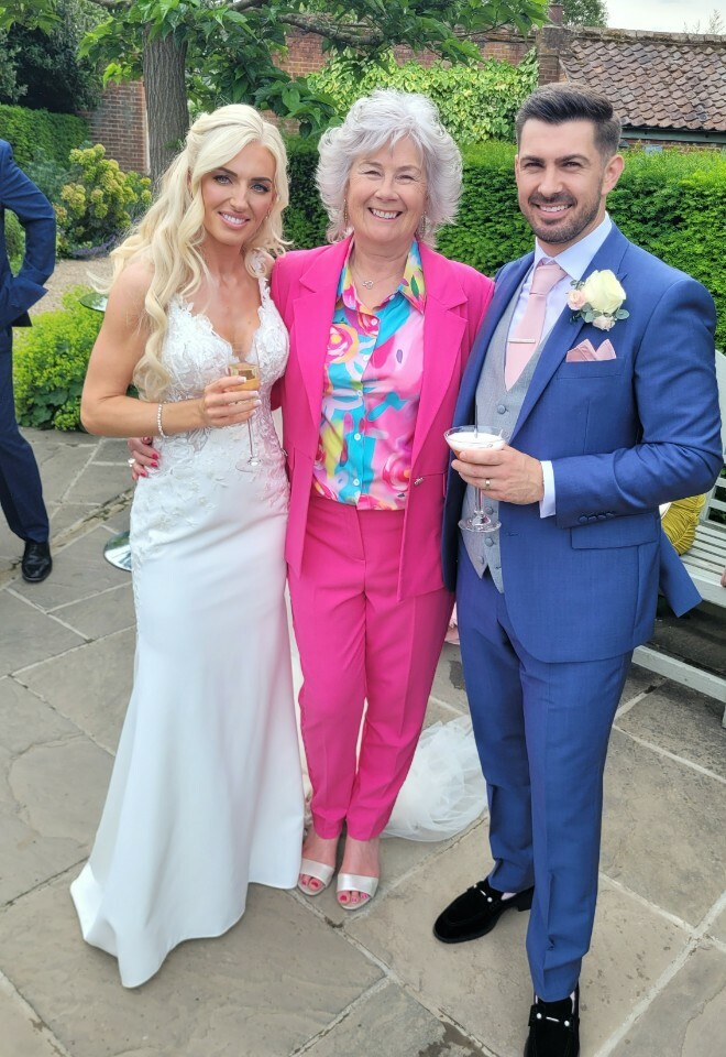 Essex Wedding celebrant dressed in a pink suit standing in between a bride and groom after their wedding ceremony