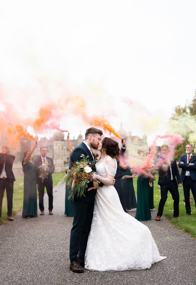 Bride & groom kissing with the bridesmaids & groomsmen holding orange & pink smoke bombs