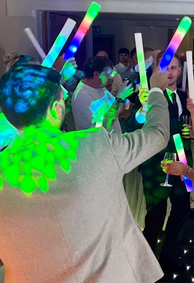 A lively wedding reception crowd dancing on a lit black dance floor, with guests waving multi-colored LED foam glow sticks under green party lights.