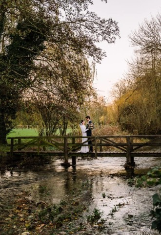 Bride and groom stand on the wooden bridge across Ampney Brook on their wedding day at Wild Thyme & Honey