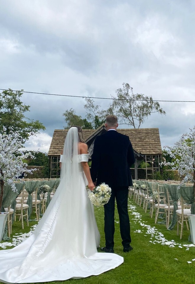 Bride and groom standing at the outdoor ceremony aisle at Great Tythe Barn, with white petals scattered on the grass, floral arrangements and chairs lined with soft green draping, facing a wooden gaze