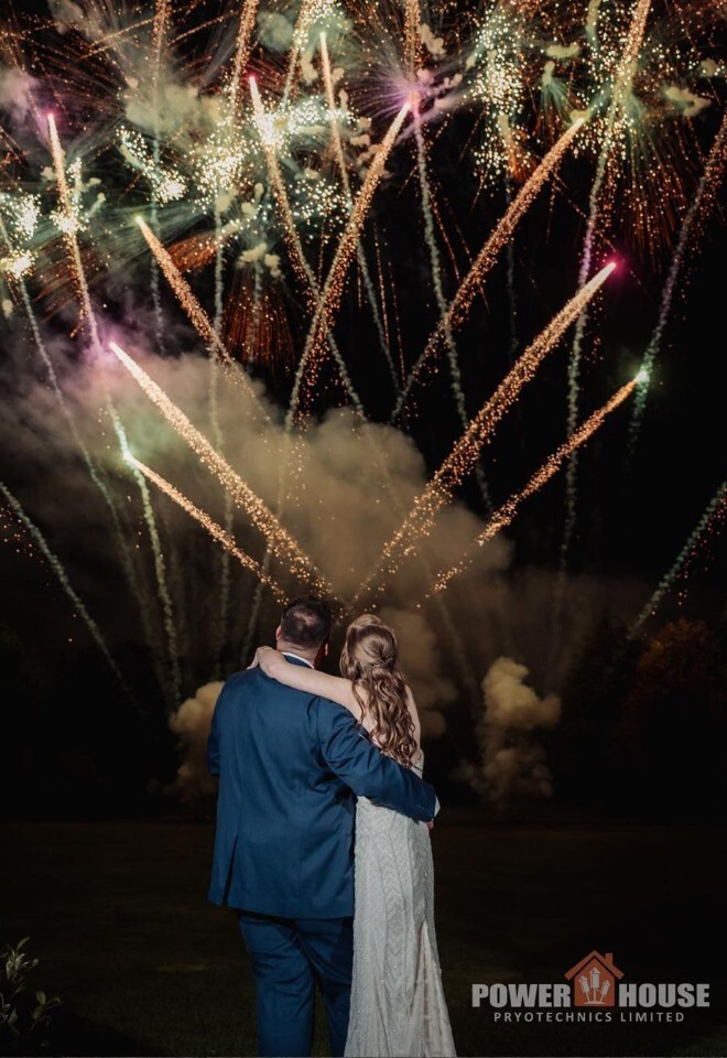 Bride and groom standing arm in arm at their wedding, viewed from behind, watching spectacular wedding fireworks lighting up the night sky above the venue at Burnham Beeches Hotel, creating a romantic