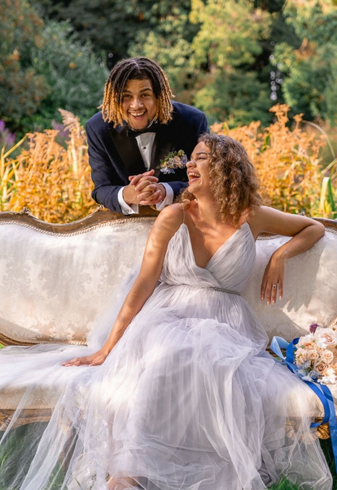 Bride and Groom laughing in the garden, bride sitting on an antique sofa, groom behind the sofa leaning on the back rest and looking at us