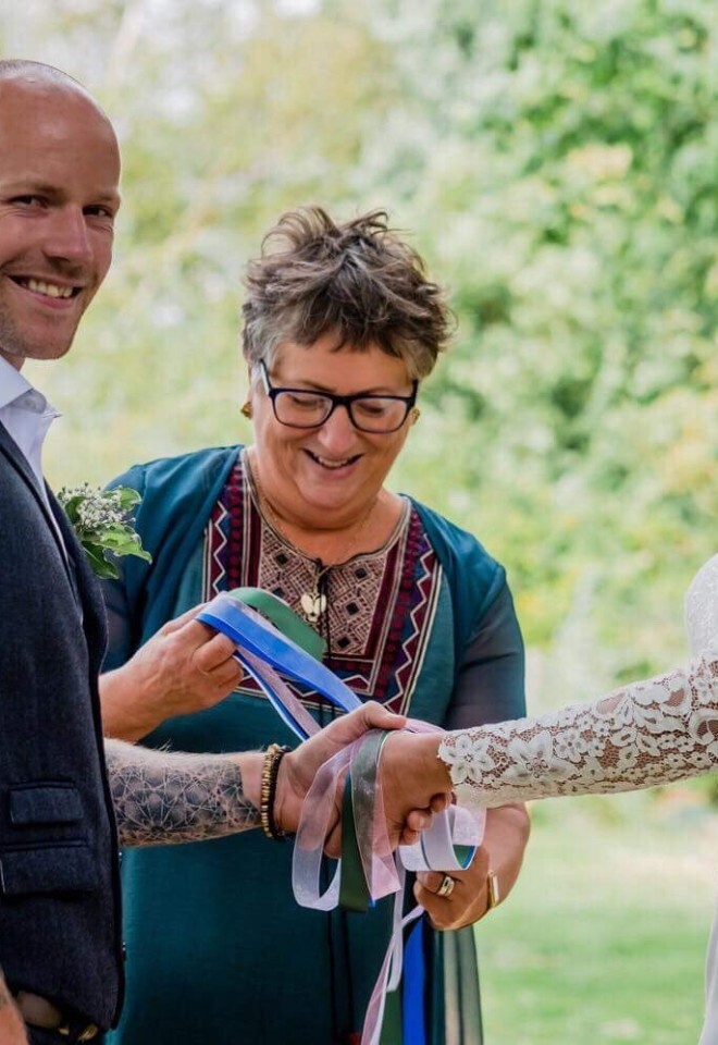 Celebrant Dawn Rees leading a handfasting ceremony outdoors with a bride and groom holding ceremonial ribbons