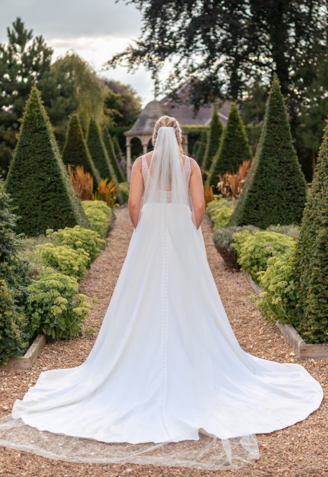 Bride in a flowing white gown and veil stands on a gravel path lined with manicured topiary trees in the gardens of Washingborough Hall Hotel.