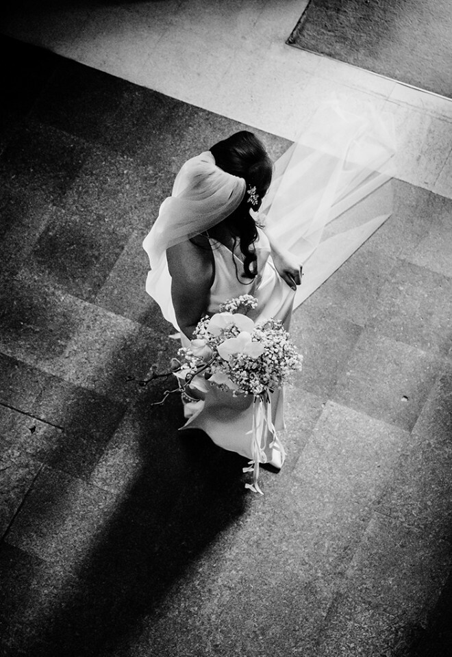 Bride walking into the ceremony, captured from above with her veil trailing behind—black and white documentary wedding photography