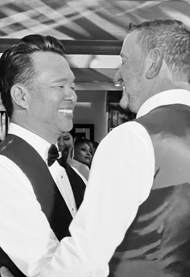 A black and white photo of a groom smiling warmly at his husband during their first dance at their wedding reception, surrounded by guests watching the moment.