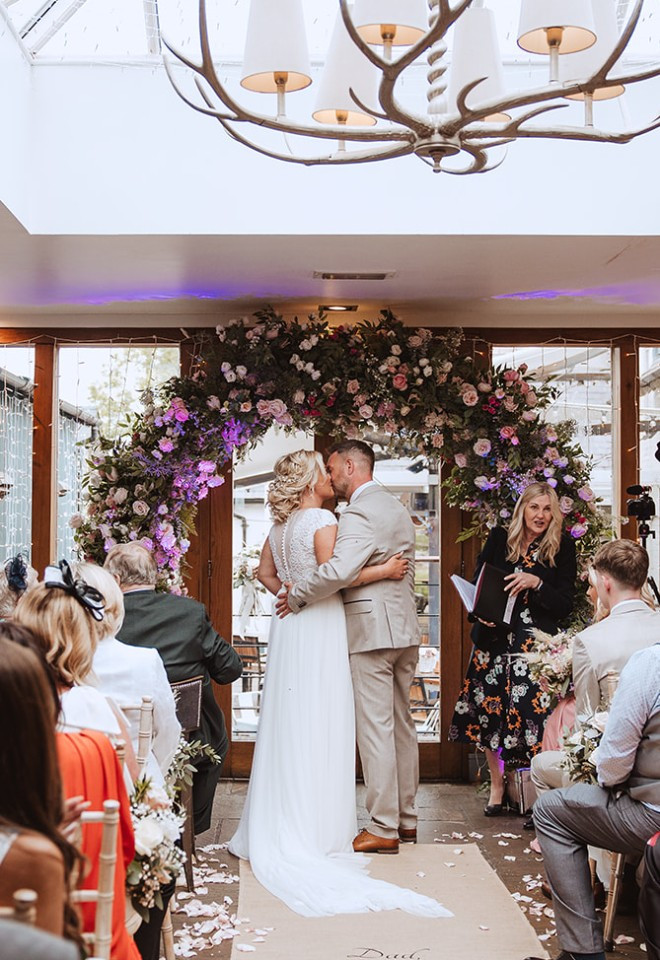 Newlyweds sharing their first kiss beneath a beautiful floral arch during their indoor ceremony at The Cock O’Barton.