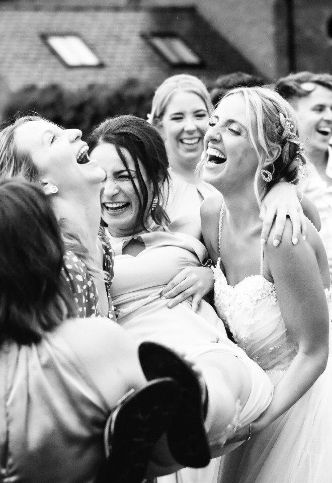 Black and white photo of people joyfully dancing and celebrating at an outdoor reception with their arms raised.