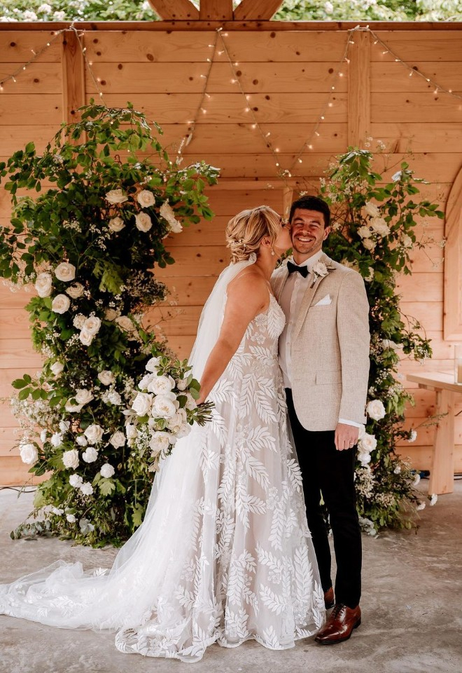 Deconstructed floral arch of white flowers and wild British foliage with bride holding white early summer bouquet at The Hidden Hive