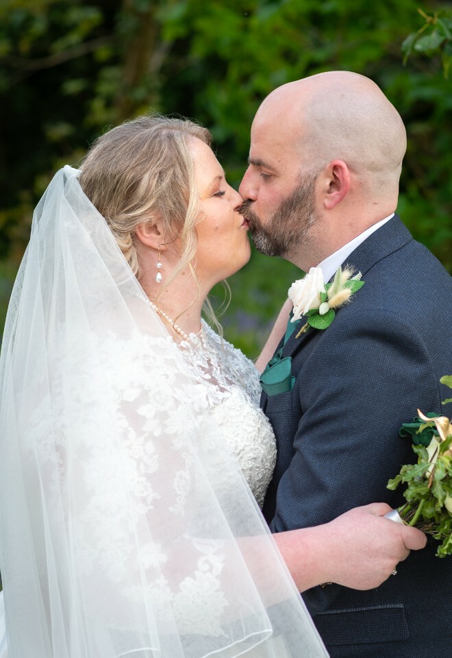 A romantic wedding kiss moment captured outdoors with greenery in the background.