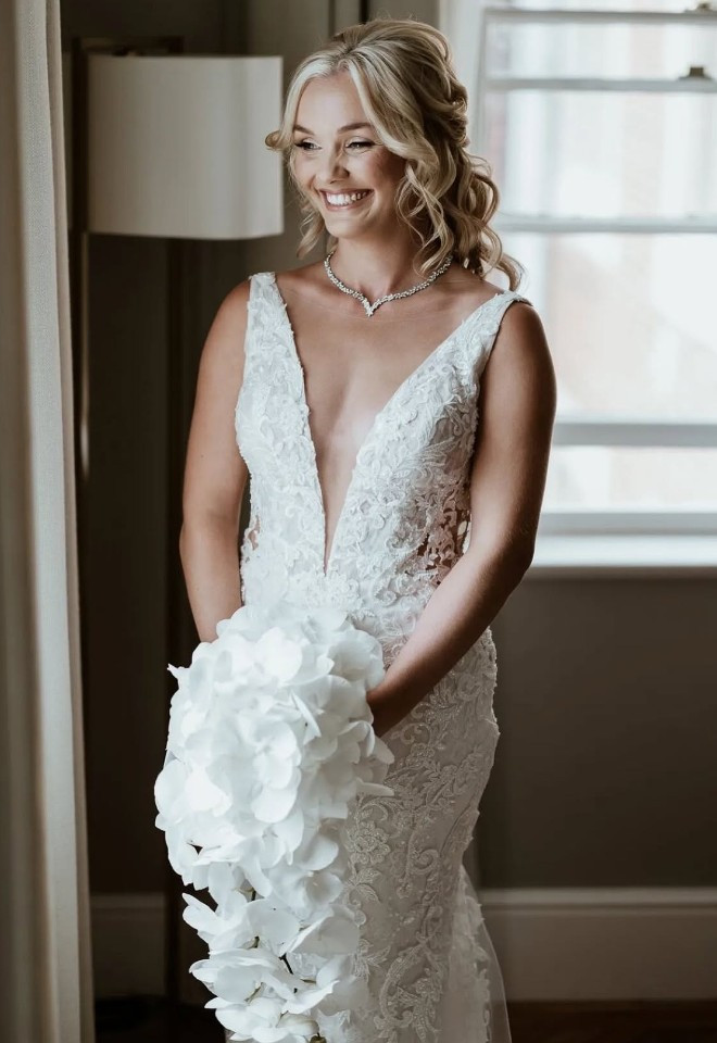 Our real bride wearing a fitted lace wedding dress by Elysee Atelie with a plunging V-neckline and delicate floral detailing, holding a cascading white bouquet, from Little London Brides in Hampshire.