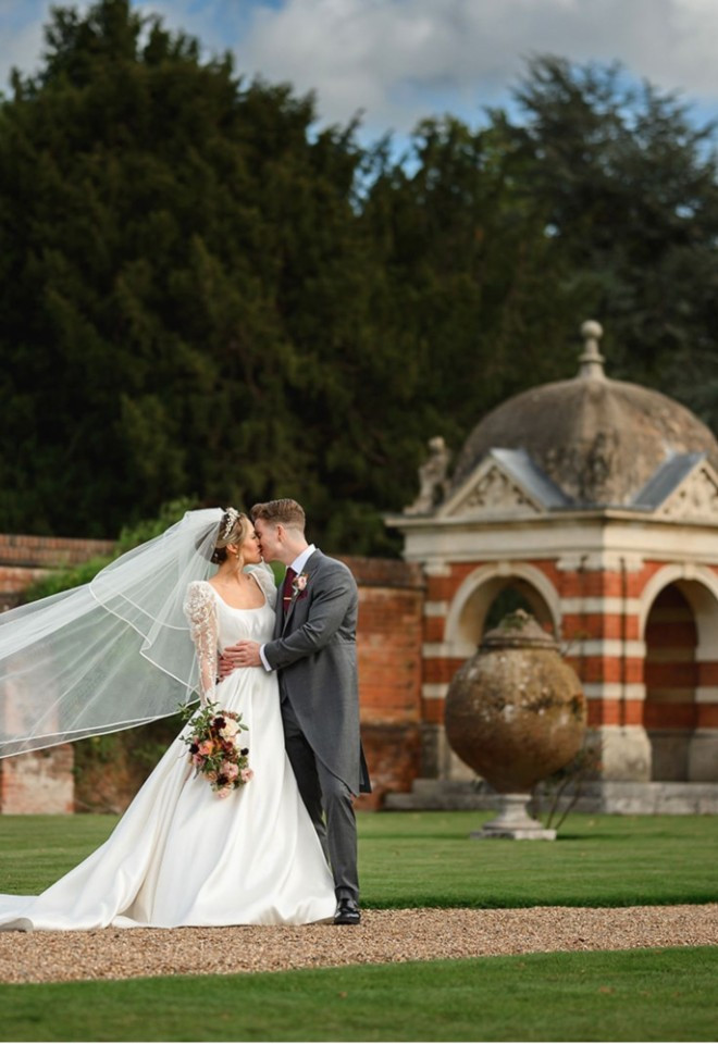 Bride and groom kissing in the gardens at North Mymms Park, with her long wedding veil floating in the wind near a classic brick pavilion.