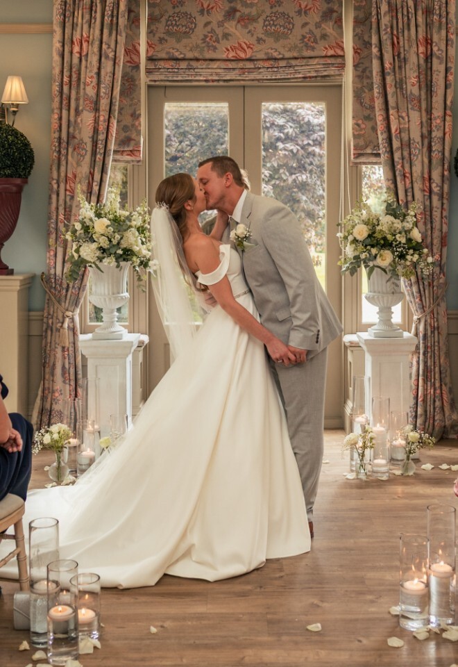 Bride and groom share their first kiss at Washingborough Hall Hotel, surrounded by guests and elegant floral arrangements in a softly lit ceremony room.