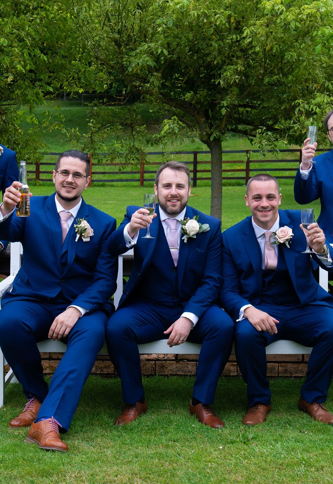 5 handsome groomsmen dressed in blue suits raise a drink to the camera whilst seated on a bench in the grounds at Sutton Bonington Hall, Leicestershire