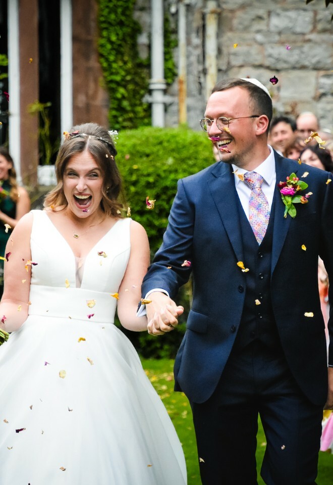 A joyful newlywed couple walks down the aisle while guests throw flower petals in celebration at an outdoor ceremony.