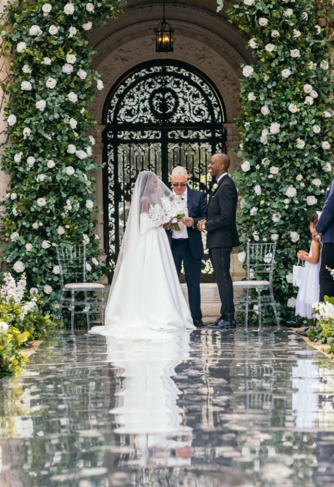 Wedding ceremony taking place in the Loggia at North Mymms Park, featuring a bride and groom at the altar framed by tall white floral arches and a reflective mirrored aisle lined with guests.