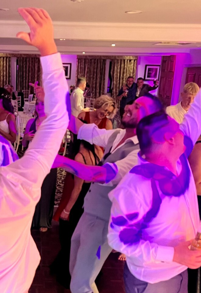 A group of wedding guests dancing enthusiastically on a dance floor, with several men in dress shirts raising their arms in the air under purple party lighting.