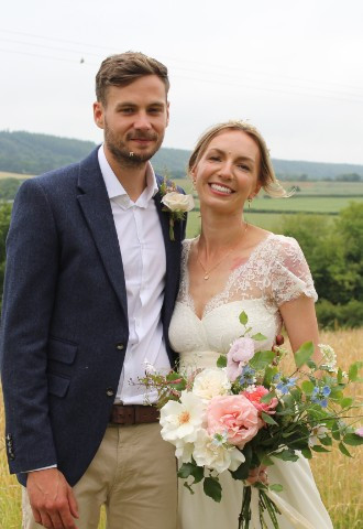 bride in lace dress holding flower bouquet with groom in blue jacket wild flower meadow background