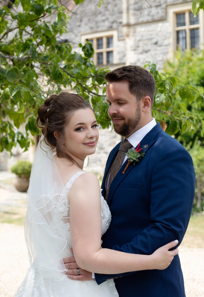 The groom lovingly looking at his stunning bride while they stand in the shade with the imposing Rhinefield House stands in the background 