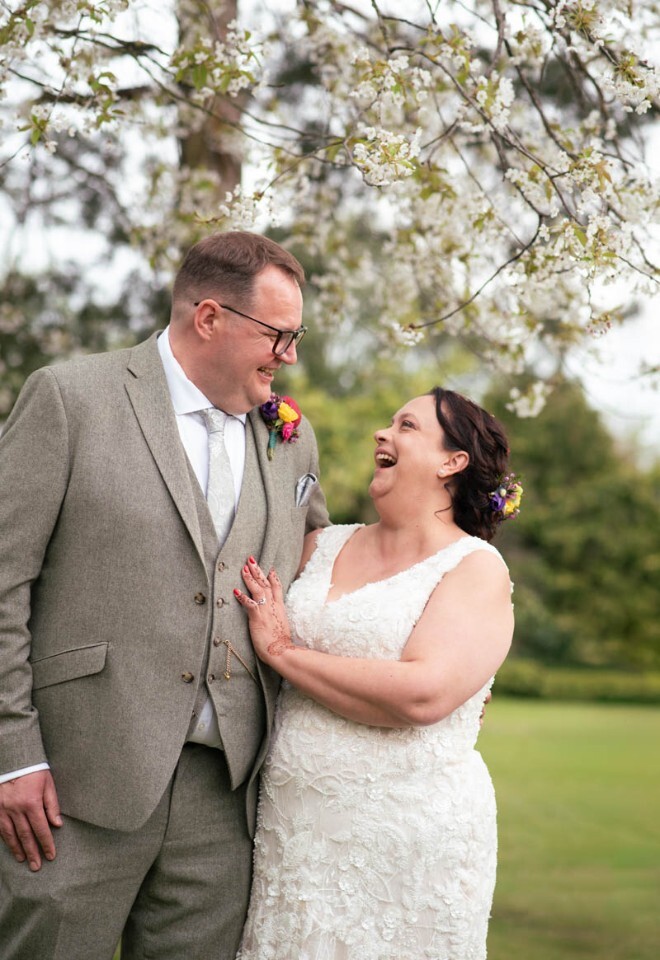 A bride and groom laugh at each other under a blossom tree. 