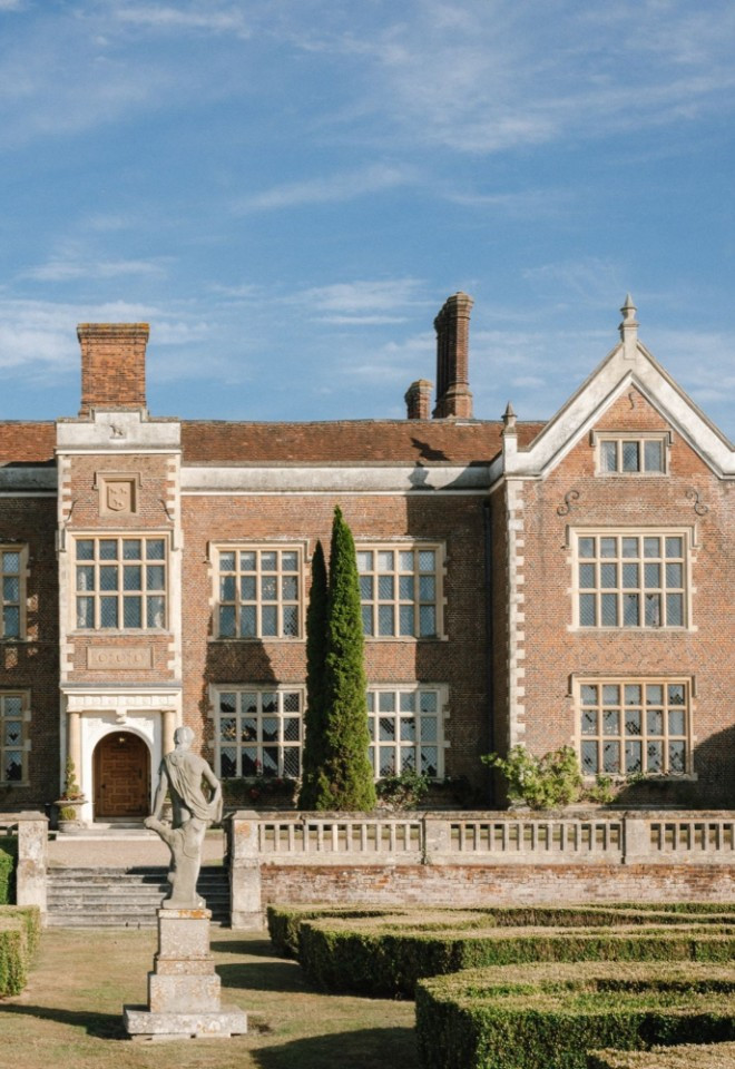 Exterior view of the historic red-brick mansion house at North Mymms Park, showcasing its symmetrical Elizabethan architecture, stone-mullioned windows, manicured hedge garden, and central statue unde