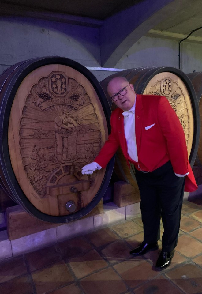 Surrey wedding toastmaster Toastmaster John in his traditional red tailcoat beside decorative wine barrels at a wedding venue, ready to host the celebrations.