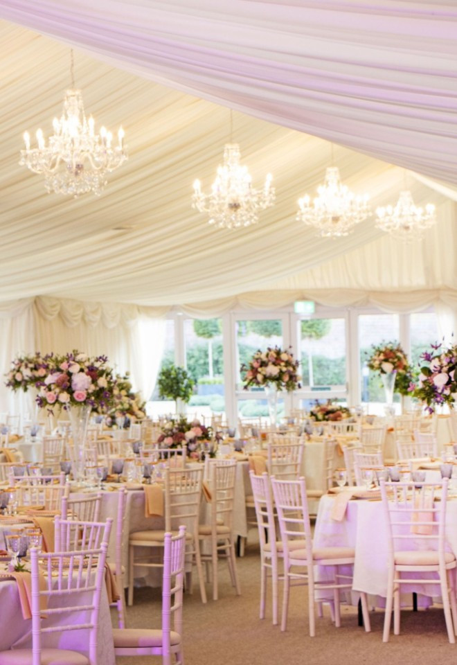 Wedding reception setup inside the Pavilion marquee at North Mymms Park, featuring round dining tables with tall floral centerpieces, white Chiavari chairs, elegant draped ceilings, and glowing crysta