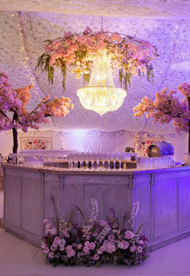 William morris linings in a luxury marquee. Pale oak bar with an arched cherry blossom trees either side and a crystal chandelier in the centre