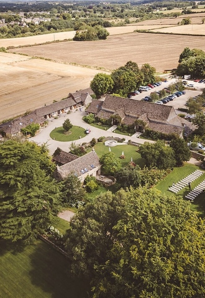 Aerial view of Great Tythe Barn surrounded by countryside fields, showing the stone barn buildings, central courtyard, gardens, outdoor ceremony setup with rows of chairs, and on-site parking.