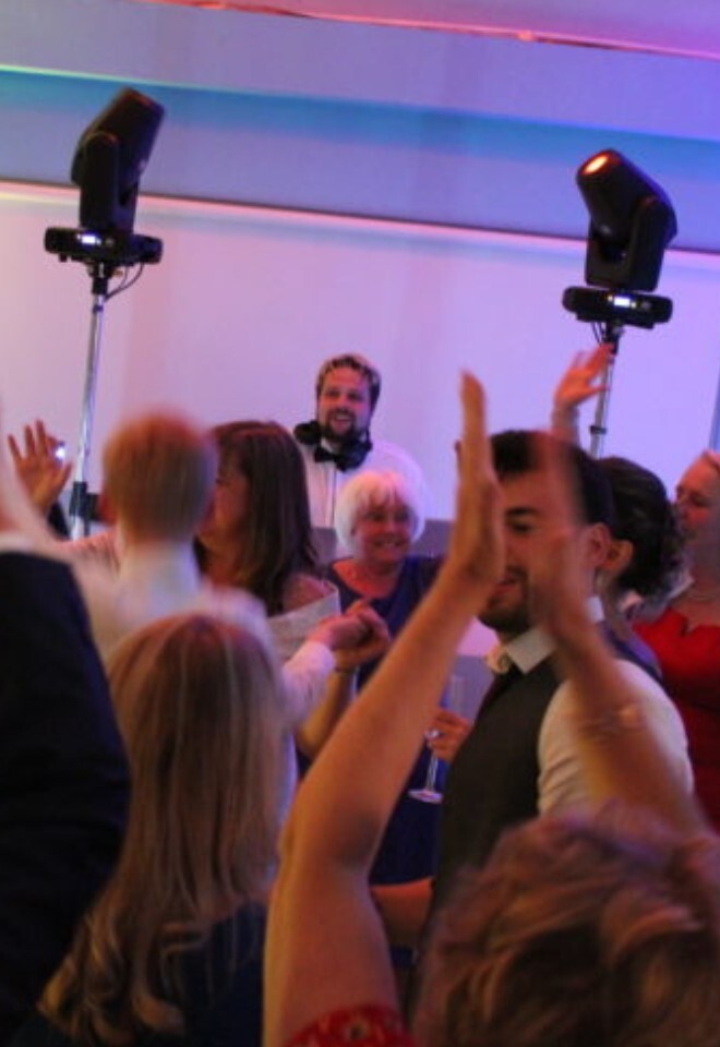 A joyful crowd dancing at a wedding reception, with several guests raising their hands in the air while a smiling DJ stands behind a booth in the background.