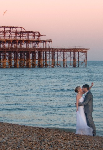 Bride and groom embrace on Brighton beach at sunset with old pier in background