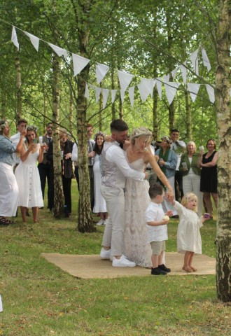 married couple dance in woodland with children surrounded by smiling wedding guests