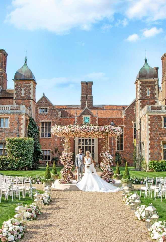 Bride and groom under a floral canopy mandap set up on the grounds of North Mymms Park, with rows of white chairs lining the gravel pathway and the red-brick mansion in the background.