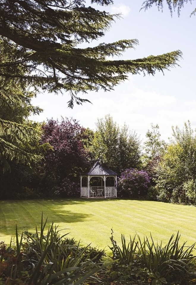 Garden lawn at Great Tythe Barn with a white gazebo set among trees and flowering greenery, framed by overhanging branches in soft sunlight.