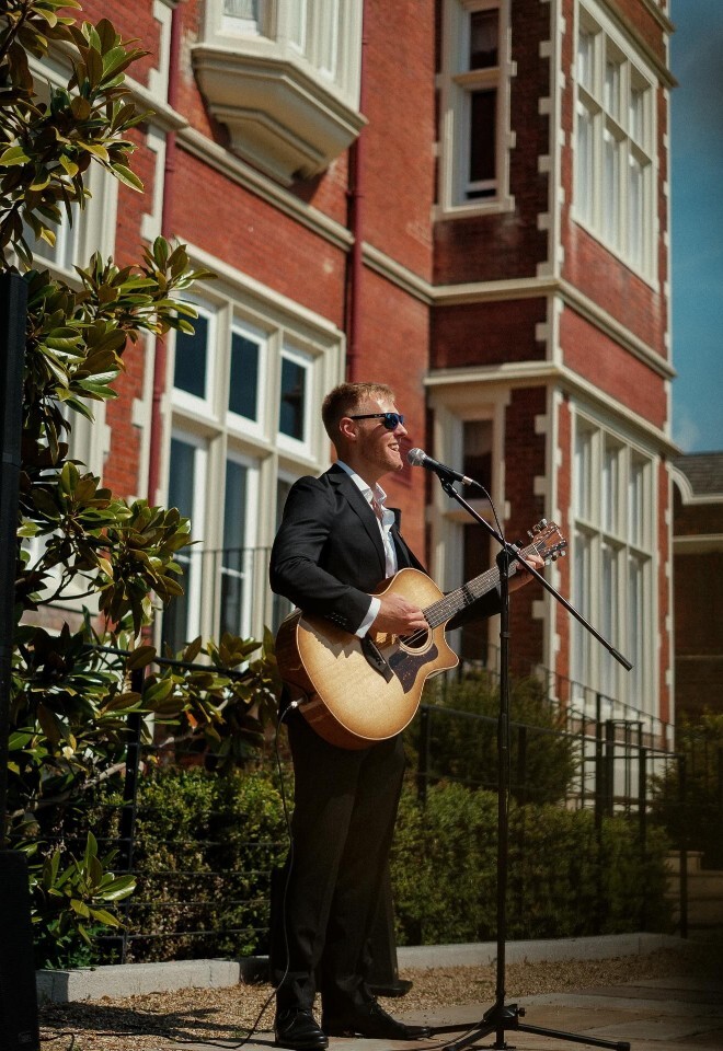 Matt Rayner, an Essex-based wedding musician, performs outdoors with his acoustic guitar in front of a grand red-brick wedding venue.