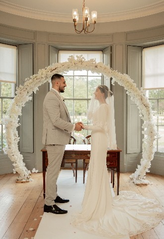 Bride & Groom stood by the registry desk with a large hoop or white flowers as a background, marrying at Valentines Mansion in Essex