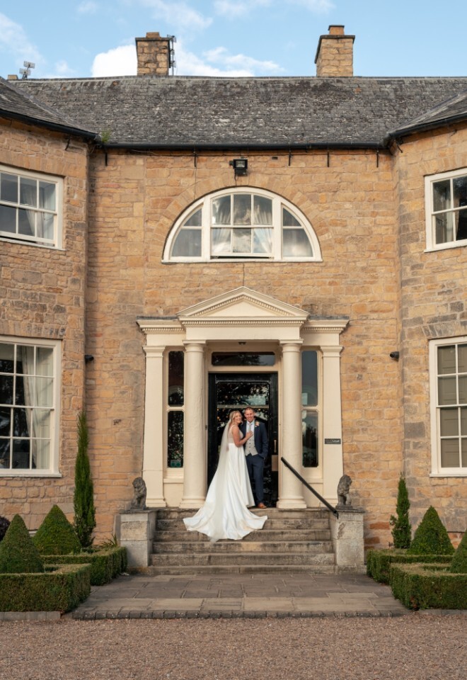 Bride and groom stand on the steps of Washingborough Hall Hotel, framed by its grand Georgian façade and manicured gardens under a clear blue sky.