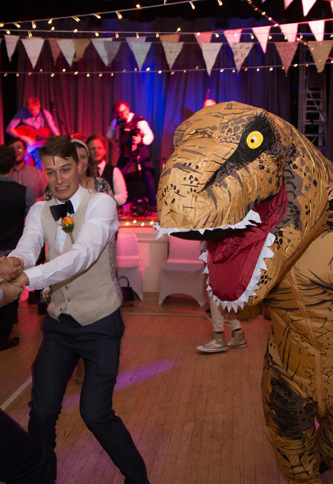 “Wedding guests dancing with a person in a dinosaur costume at a reception at Horton and Chalbury Village Hall in Dorset photographed by Katy Brothers Wedding Photography”